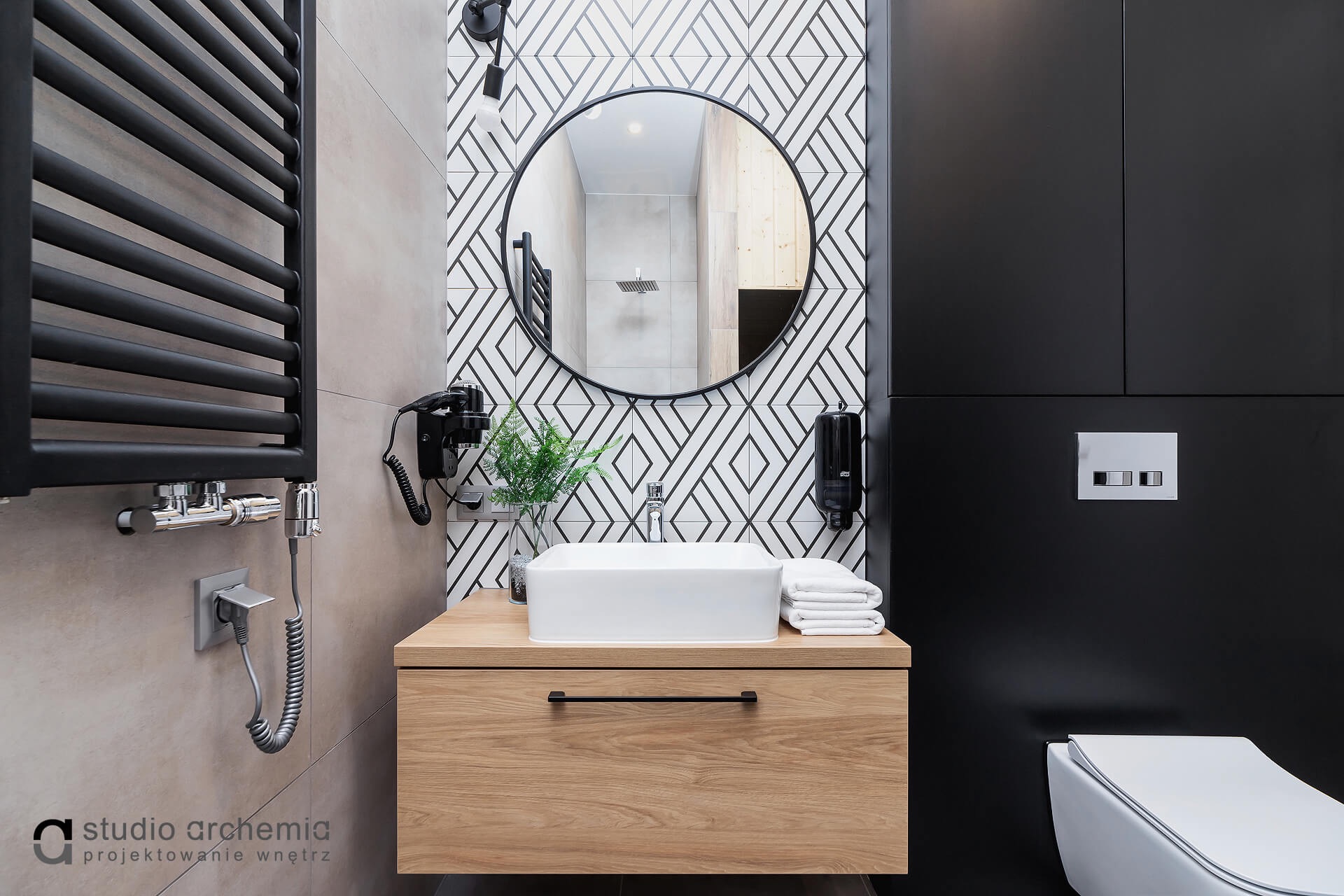 Nowoczesna łazienka z drewnianą szafką pod umywalką, okrągłym lustrem i geometryczną mozaiką na ścianie, uzupełniona czarnym grzejnikiem drabinkowym i zabudową.