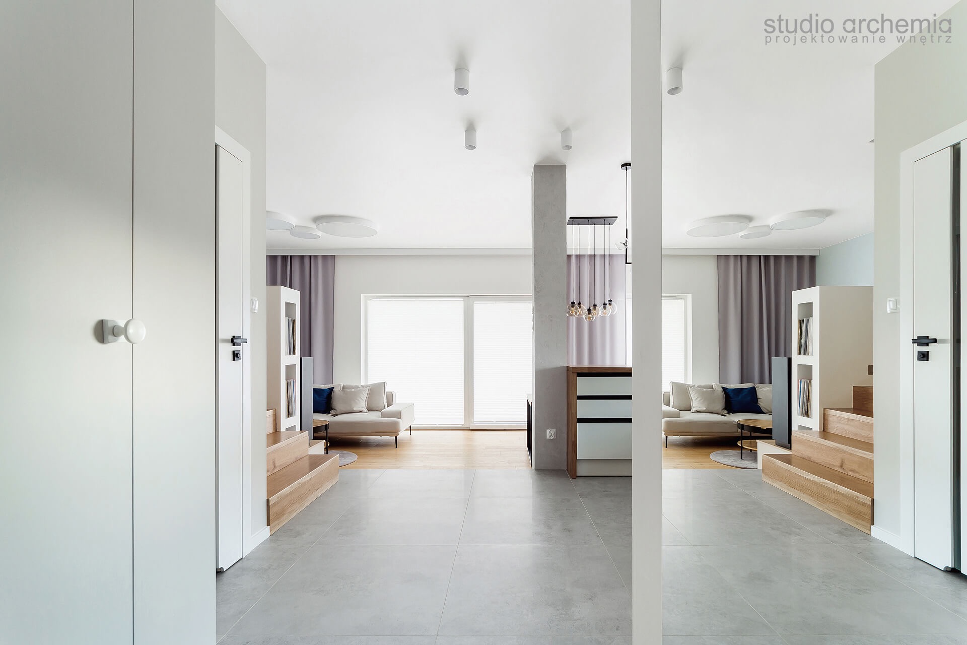 Nowoczesne wnętrze z szarą podłogą, białymi ścianami, drewnianymi schodkami i sofą z niebieskimi poduszkami widziane z perspektywy wejścia, z logo 'studio archemia' w prawym górnym rogu.