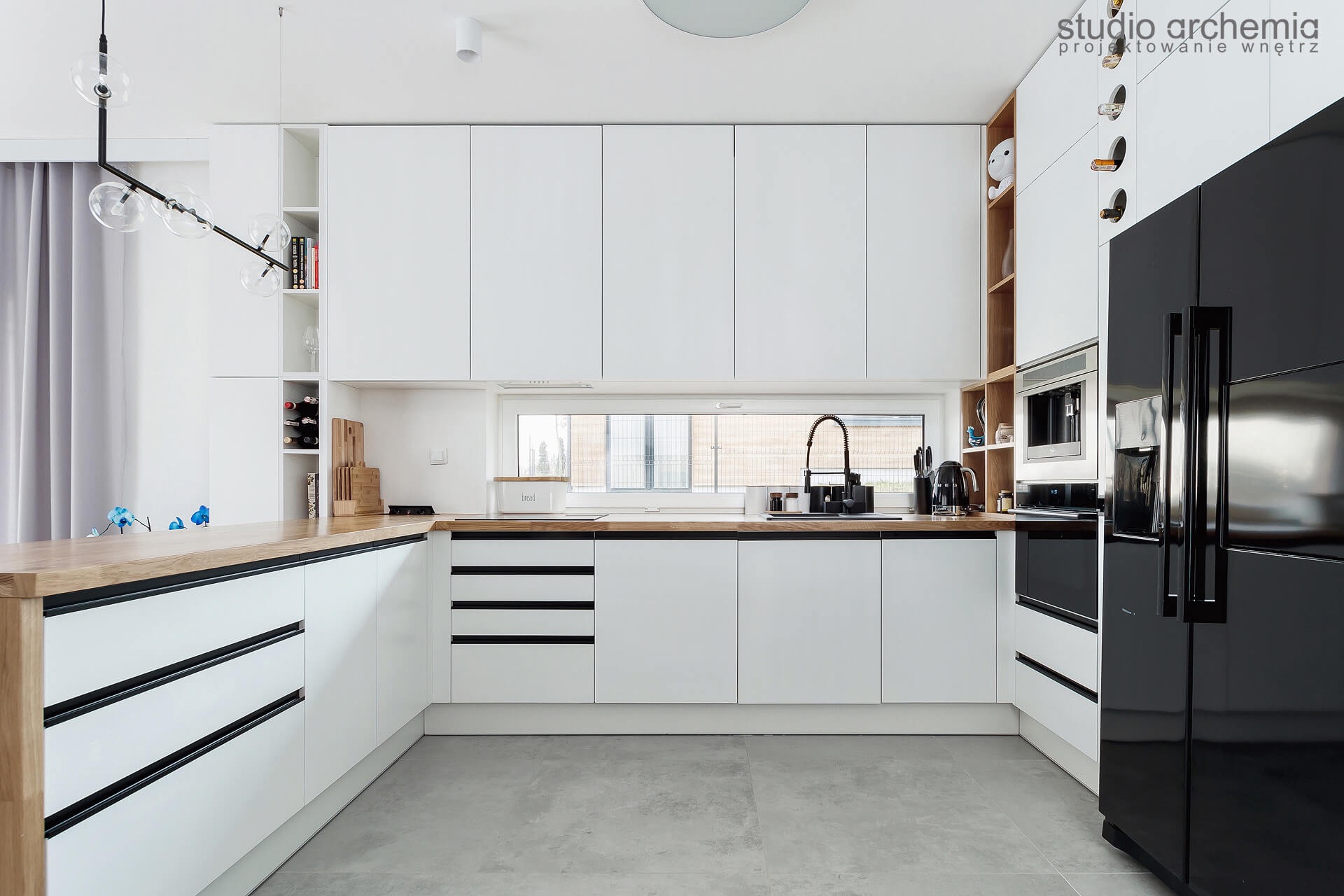 Nowoczesna kuchnia z białymi szafkami i drewnianym blatem, czarną lodówką i designerską lampą wiszącą, widok na okno z czarną armaturą.