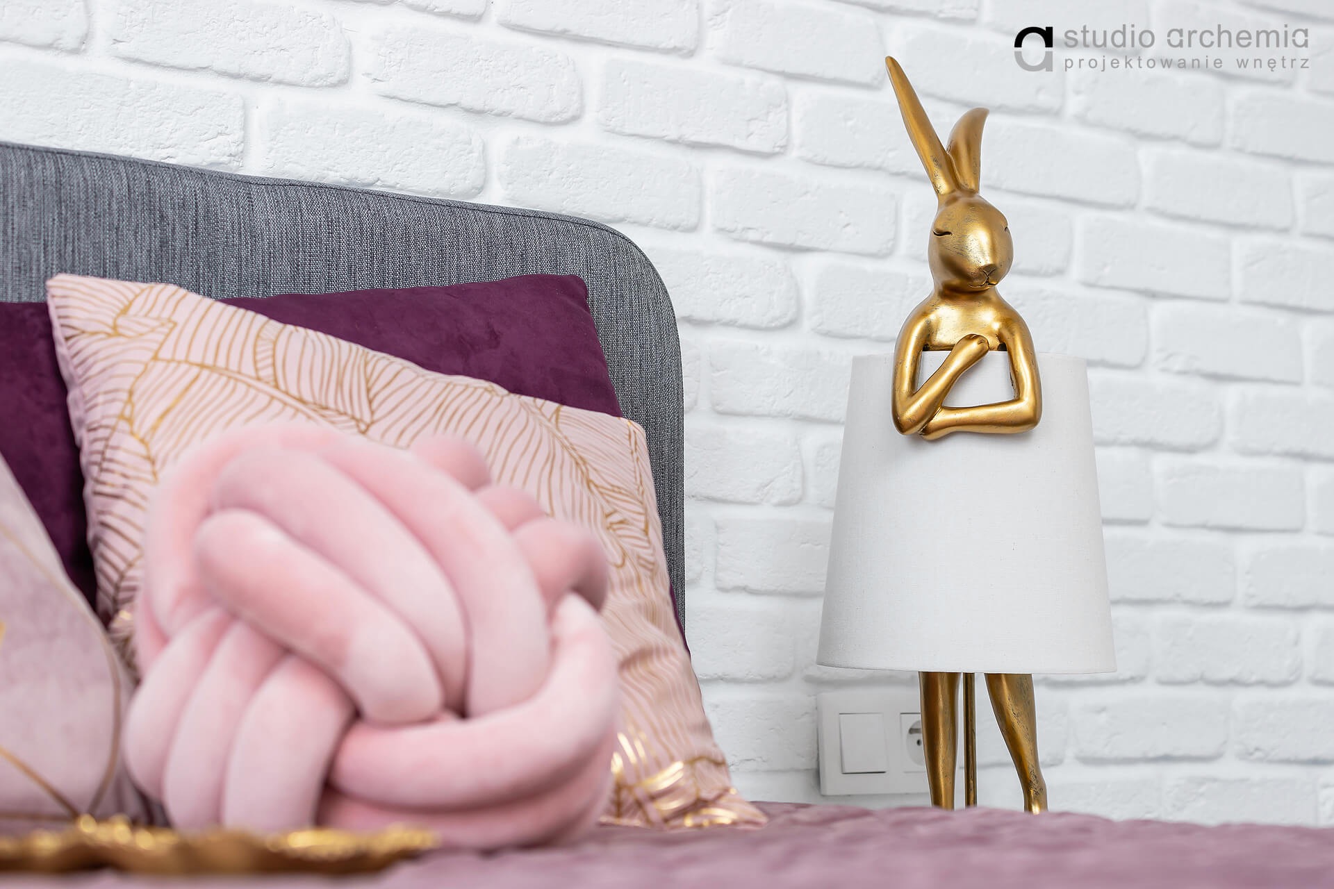 Sypialnia z dekoracyjną lampką nocną w kształcie złotego królika na tle białej ściany z cegieł, obok poduszki w kształcie węzła i poduszki dekoracyjnej z motywem liści.