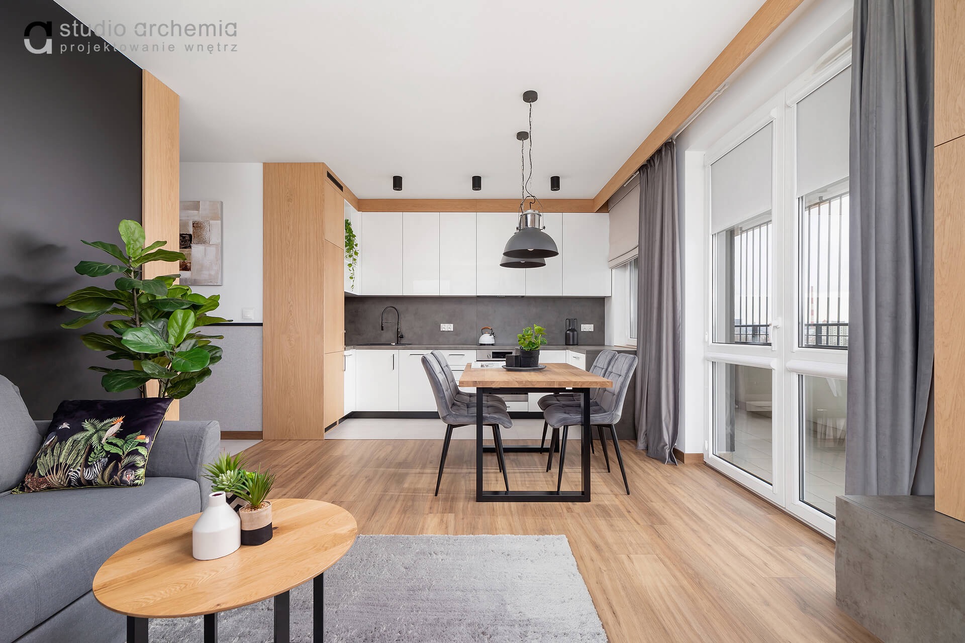 Jasne, nowoczesne wnętrze mieszkania z drewnianą podłogą, szarą sofą z poduszką w tropikalne wzory, okrągłym stolikiem kawowym i stołem jadalnym z szarymi krzesłami, oświetlonym przez wiszące lampy...