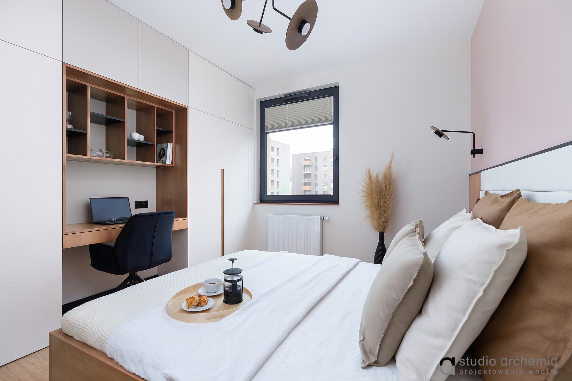 Nowoczesna sypialnia z biurkiem w zabudowie, łóżkiem z tacą śniadaniową i dekoracyjną trawą pampasową w czarnym wazonie.