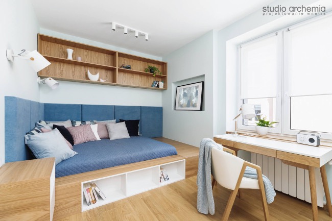 Pokój z podwyższonym łóżkiem z niebieskim zagłówkiem i kołdrą, drewnianym regałem nad nim, biurkiem z krzesłem przy oknie i jasną podłogą.