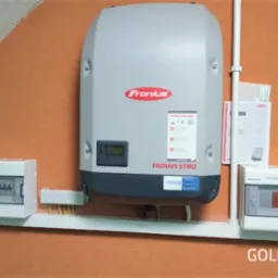 Zamontowany inwerter Fronius Symo na pomarańczowej ścianie, z widocznymi skrzynkami bezpiecznikowymi i okablowaniem, wraz z dokumentacją techniczną w tle.