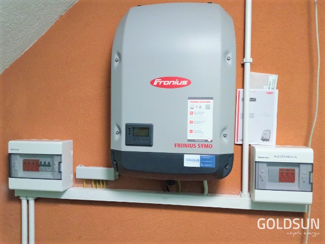 Zamontowany inwerter Fronius Symo na pomarańczowej ścianie, z widocznymi skrzynkami bezpiecznikowymi i okablowaniem, wraz z dokumentacją techniczną w tle.