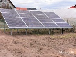 Instalacja paneli słonecznych na konstrukcji wsporczej, widoczna z poziomu gruntu, z domem i tunelem foliowym w tle.