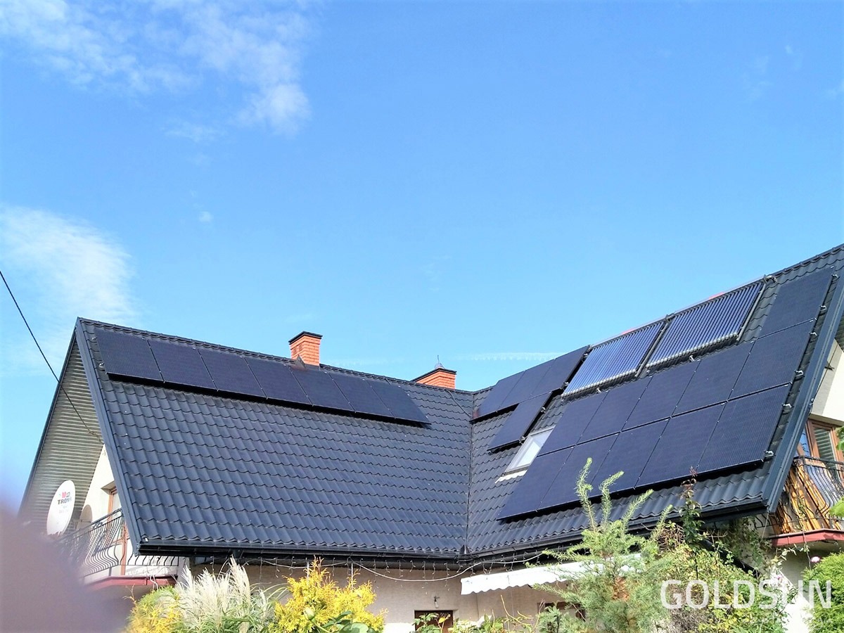 Dom jednorodzinny z zainstalowanymi panelami fotowoltaicznymi i kolektorami słonecznymi na dachu z czarnej dachówki, widoczne krzewy w ogrodzie.