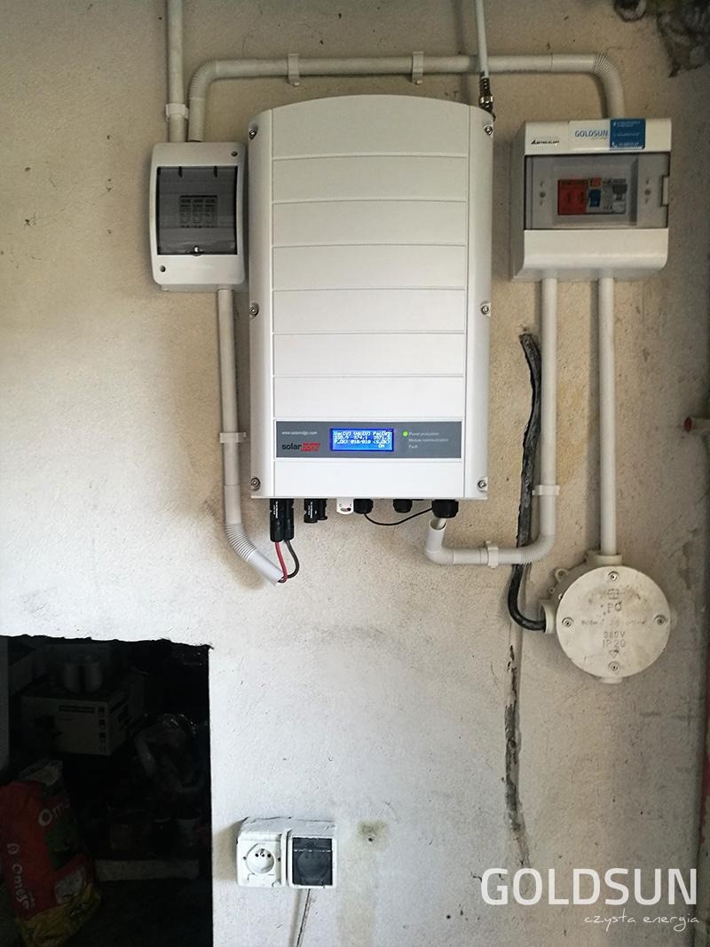 Biały falownik fotowoltaiczny SolarEdge zamontowany na surowej, szarej ścianie wewnątrz budynku, z widocznymi przewodami, skrzynką bezpiecznikową i gniazdkiem elektrycznym poniżej, oraz logiem...