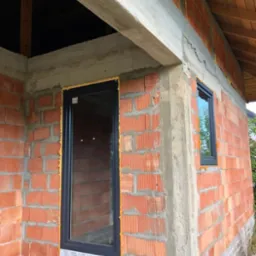 Montaż okien w surowym budynku z czerwonej cegły, widoczne czarne ramy okienne i pianka montażowa.