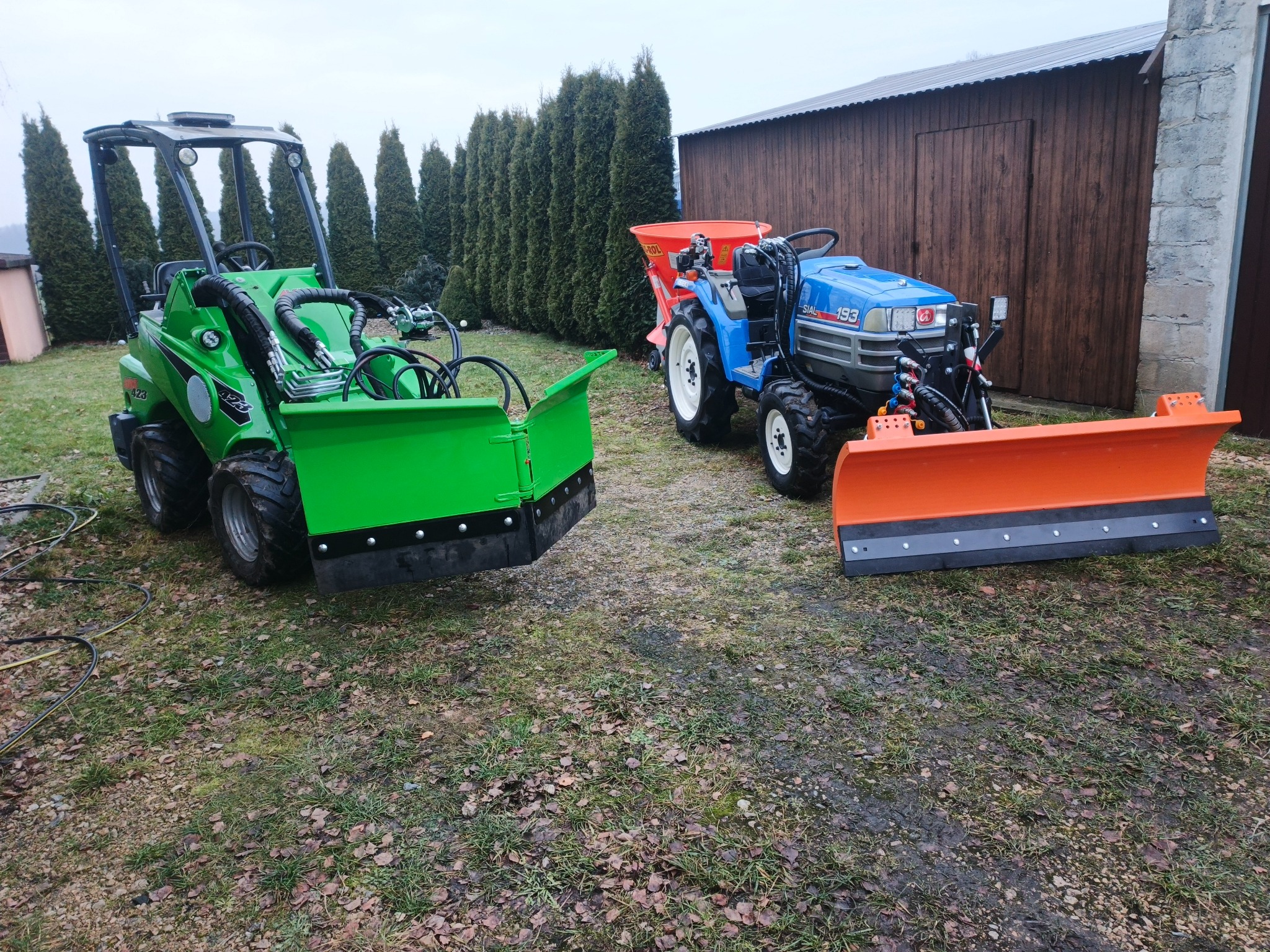 Dwa traktory: zielony z przodu z pługiem i niebieski z rozsiewaczem na tle żywopłotu i drewnianego budynku. Sprzęt do pielęgnacji trawnika i odśnieżania.