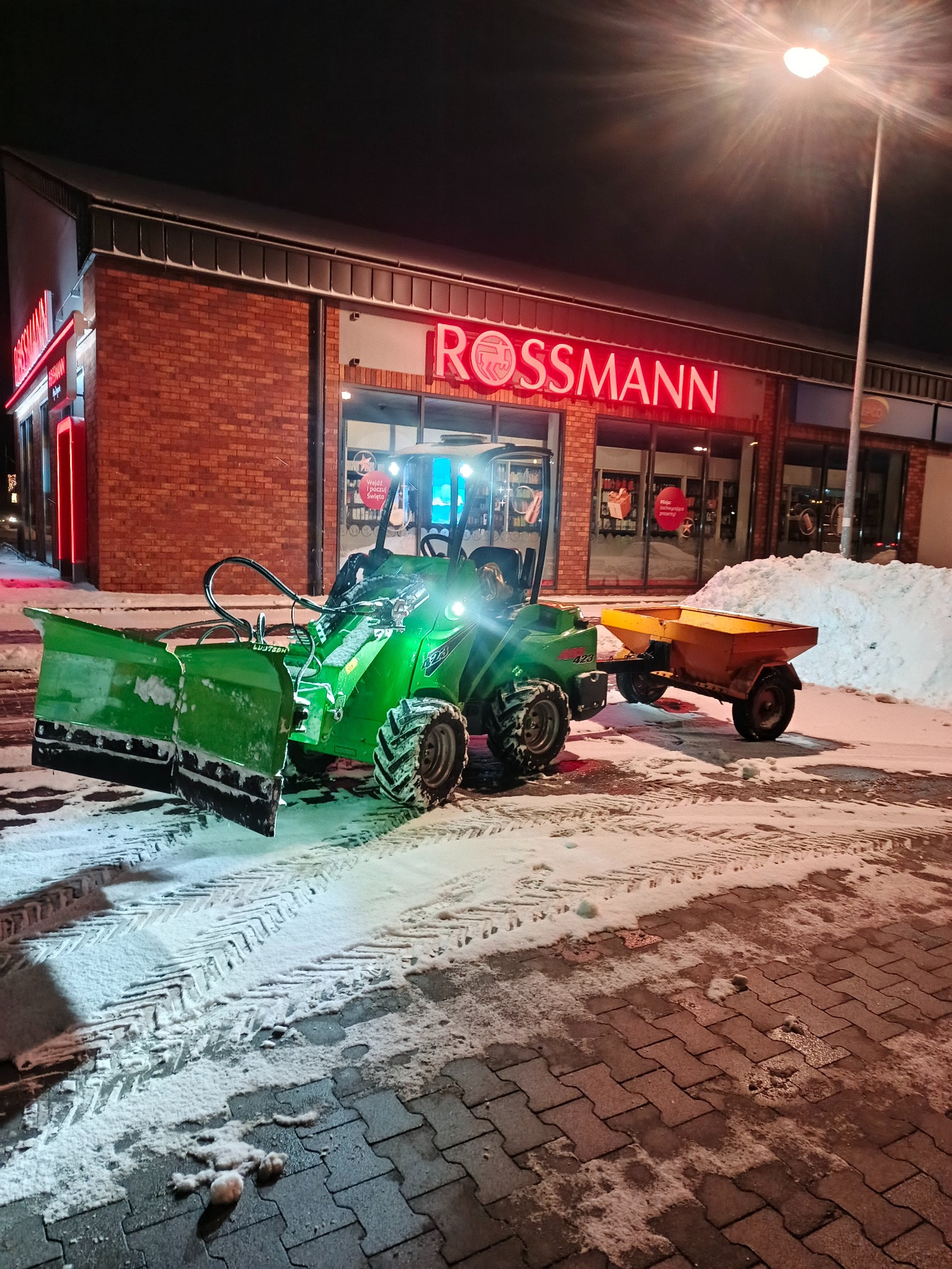 Zielony traktor z pługiem i przyczepą odśnieża parking przed sklepem Rossmann nocą. Ślady opon na śniegu, oświetlenie uliczne.
