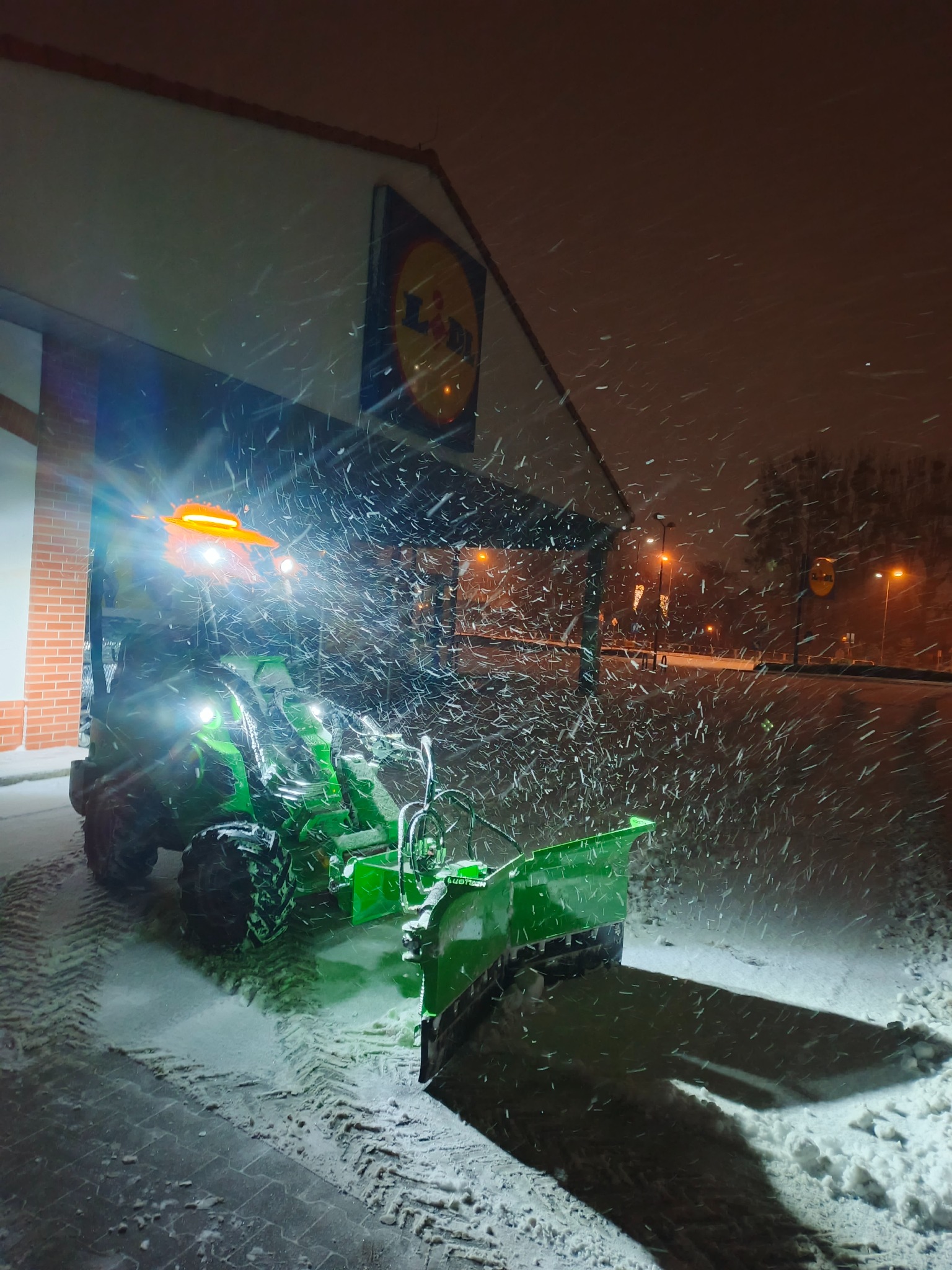 Zielony traktor z pługiem odśnieżającym, oświetlony, usuwa śnieg na tle budynku z logo Lidl podczas intensywnych opadów śniegu nocą.