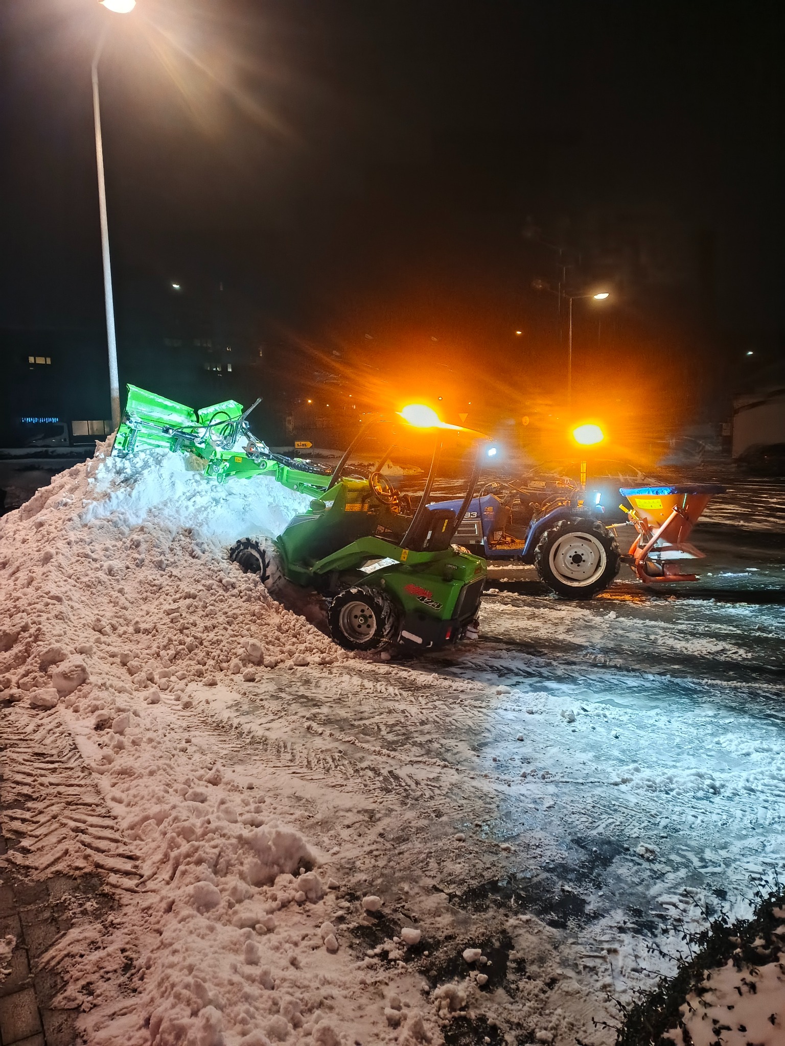 Nocne odśnieżanie parkingu: zielony traktor z frezem wirnikowym i niebieski z posypywarką, oświetlone lampami ulicznymi, ślady opon na śniegu.