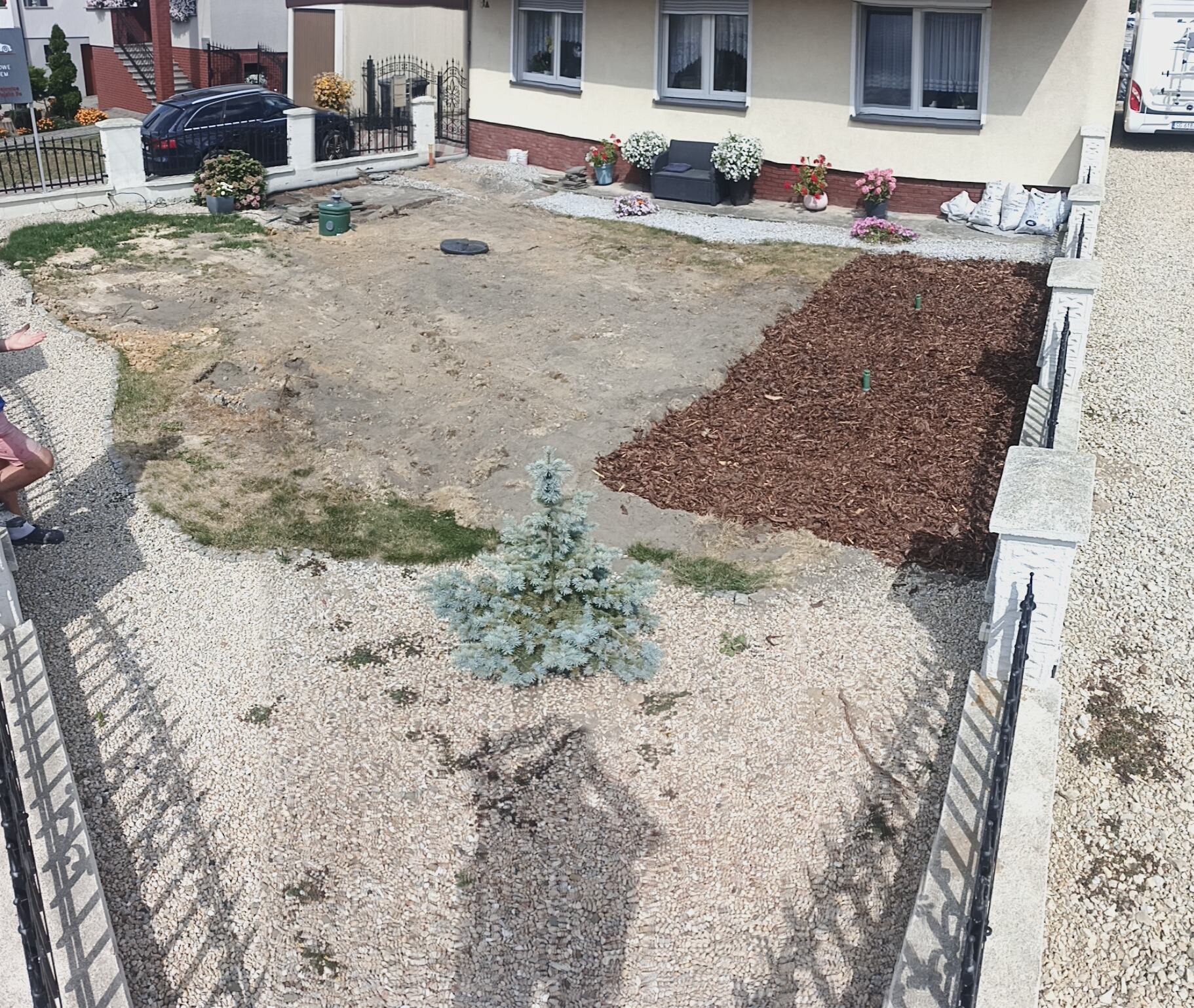 Widok z góry na ogród w trakcie aranżacji: jasne kamienie, kora, posadzona choinka, przygotowany teren pod trawnik. Częściowo widoczny budynek i ogrodzenie.