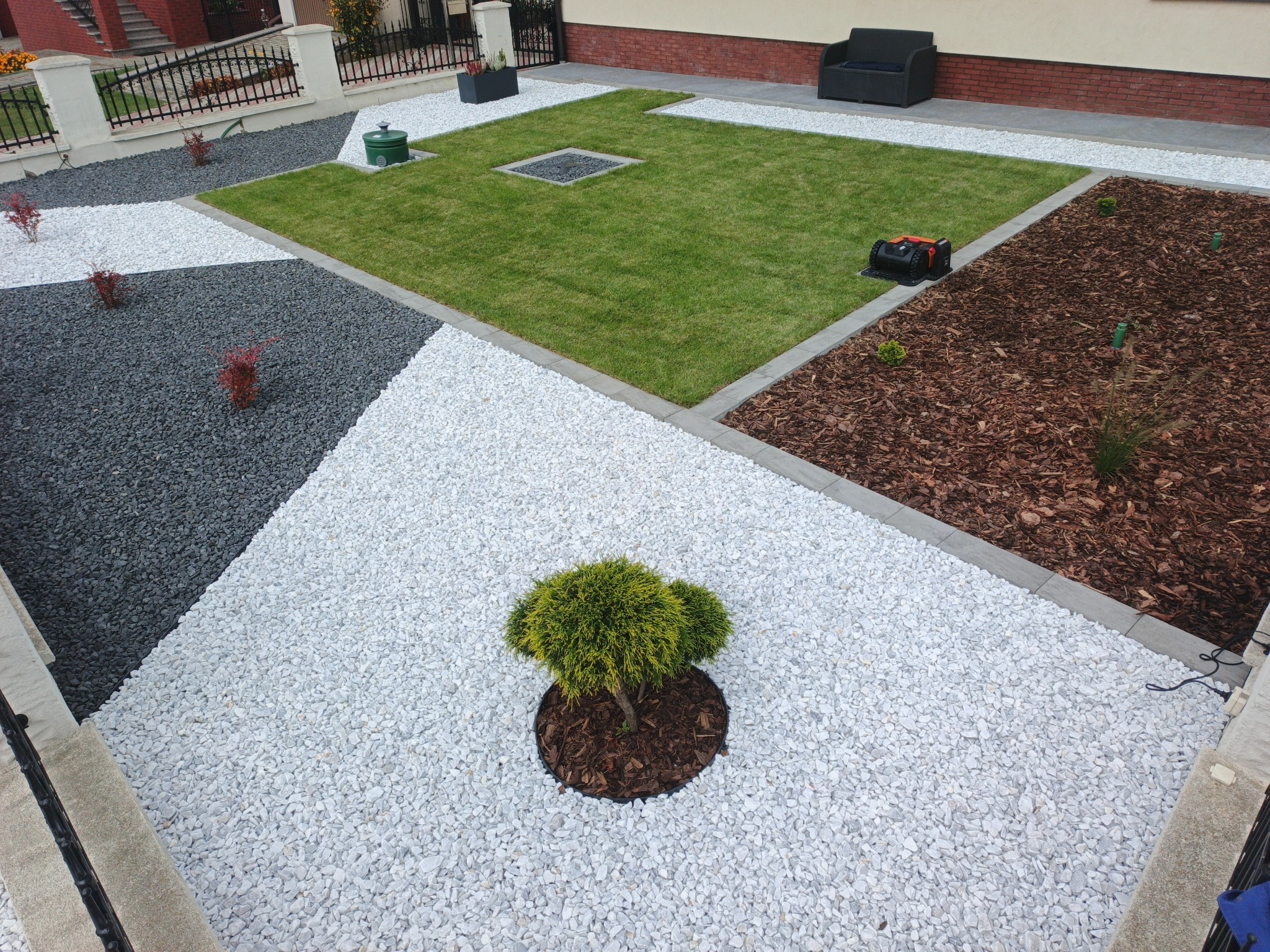 Nowoczesny ogród z geometrycznym układem trawnika, białych i szarych kamieni, kory oraz roślin. Robot koszący na korze. Widok z góry.