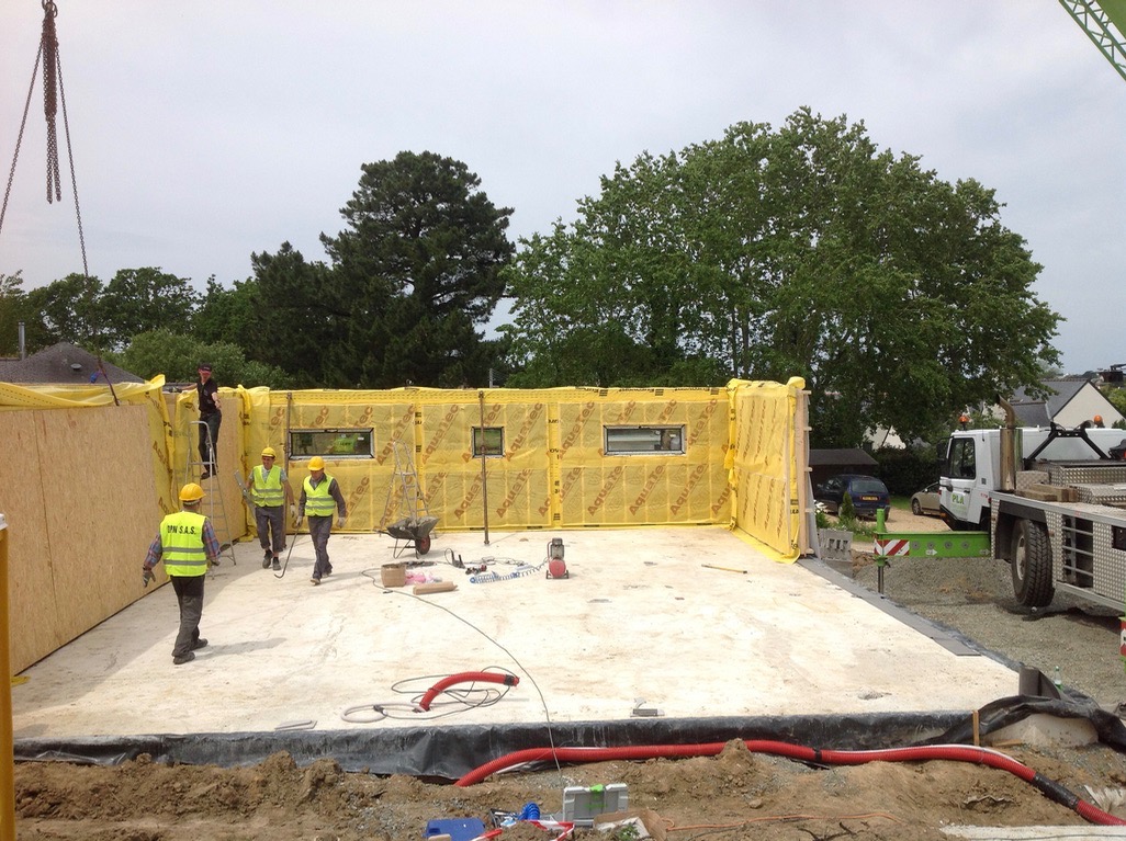 Montaż segmentów domu szkieletowego z widoczną izolacją Aquatec, pracownicy w kamizelkach odblaskowych, żuraw i częściowo widoczny samochód ciężarowy na tle zieleni.