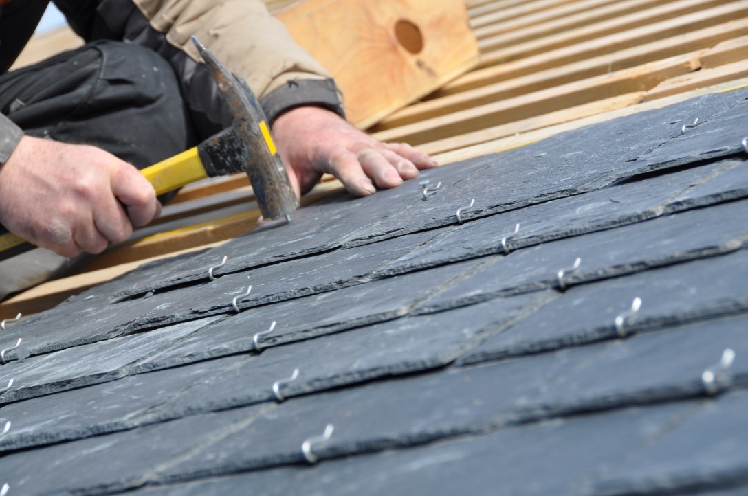 Ręczne układanie ciemnych, łupkowych dachówek na drewnianej konstrukcji dachu, zabezpieczonych metalowymi klamrami, przy użyciu młotka z żółtym trzonkiem.
