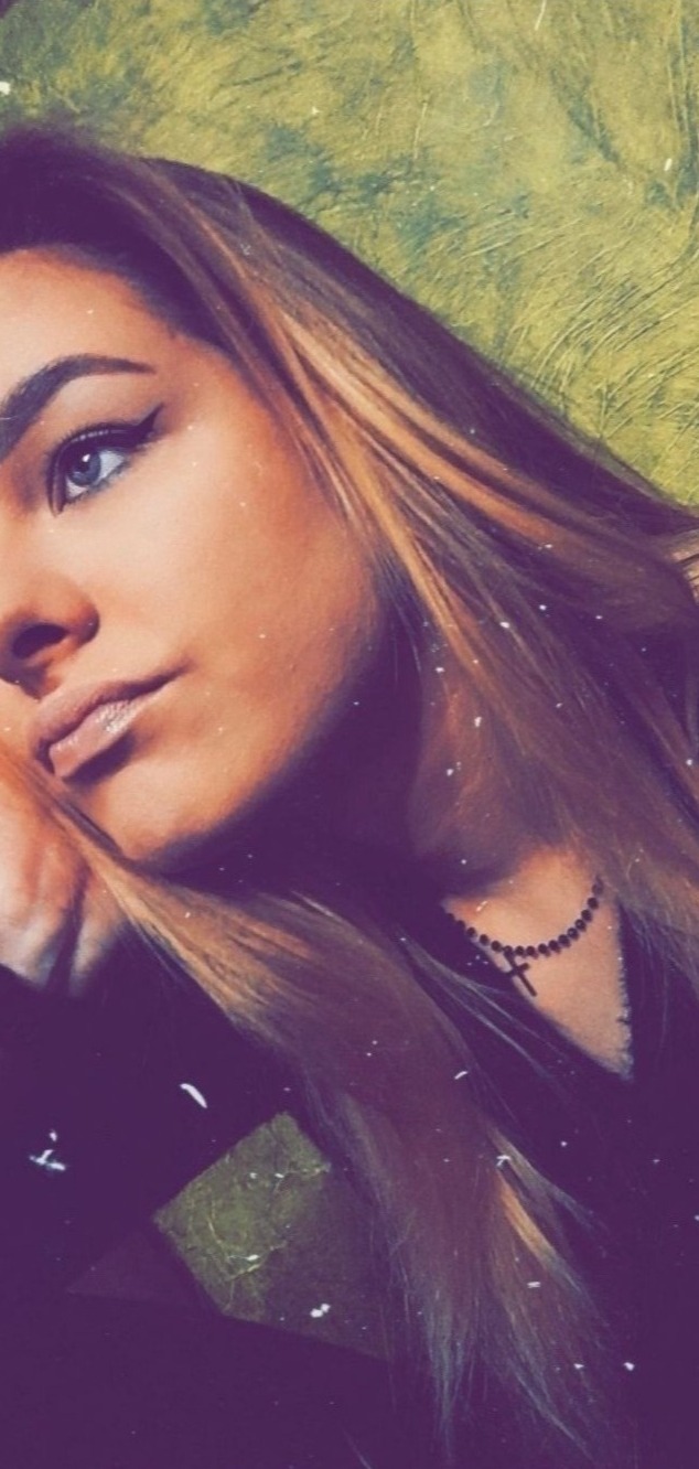 Portret kobiety z długimi blond włosami, czarną kreską na powiece i naszyjnikiem z krzyżykiem, oparta twarzą na dłoni na tle zielonej ściany.