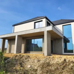 Surowy stan budynku jednorodzinnego z płaskim dachem i dużymi oknami, widoczny na tle błękitnego nieba, z odsłoniętym gruntem wokół fundamentów.