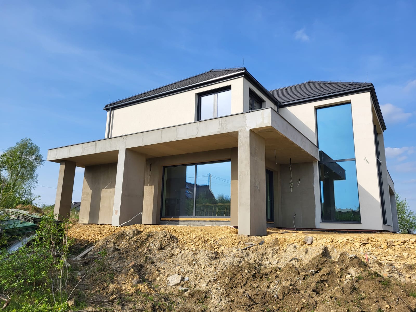 Surowy stan budynku jednorodzinnego z płaskim dachem i dużymi oknami, widoczny na tle błękitnego nieba, z odsłoniętym gruntem wokół fundamentów.