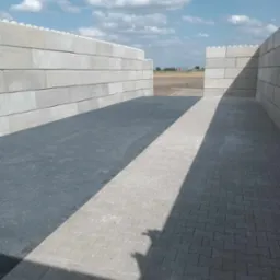 Świeżo ułożona, szara kostka brukowa w przestrzeni ograniczonej ścianami z szarych betonowych bloków, z widokiem na pole i błękitne niebo w tle.