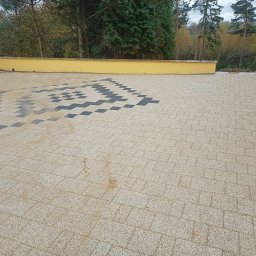 Dawid Szulik - Ułożona kostka brukowa w geometryczny wzór z ciemnych i jasnych kostek na podjeździe, widoczne żółte ściany budynku i drzewa w tle.