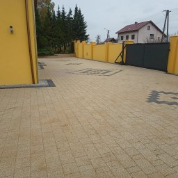 Dawid Szulik - Beżowa kostka brukowa ułożona we wzór z ciemnymi akcentami, otoczona żółtym budynkiem i ogrodzeniem, z czarną bramą w tle.