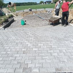 Dawid Szulik - Ułożona szara kostka brukowa na posesji, w tle pracownicy z taczką, betoniarka i widok na zielone pole oraz zabudowania.