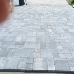 Świeżo ułożona, szara kostka brukowa w różnych rozmiarach, tworząca wzór na posesji, z widocznym fragmentem ogrodzenia i elementami krajobrazu w tle.