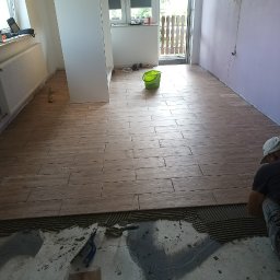 Dawid Szulik - Układanie podłogi z płytek imitujących drewno w jasnym pomieszczeniu, widoczne narzędzia glazurnicze i częściowo ułożona podłoga, w tle widok na balkon.