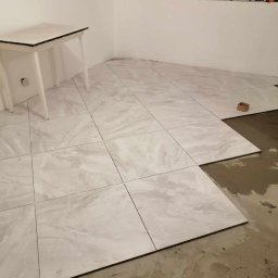 Dawid Szulik - Wnętrze pokoju w trakcie układania jasnych, marmurkowych płytek podłogowych; widoczny fragment podłogi z nieułożonymi płytkami i śladami kleju oraz biały stolik pomocniczy w rogu pomieszczenia.
