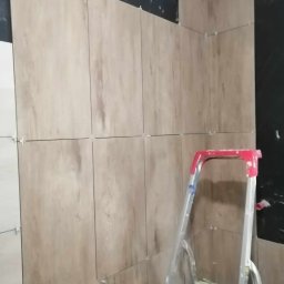 Dawid Szulik - Ściana w trakcie wykańczania płytkami imitującymi drewno, widoczna drabina malarska i niezakończone prace na górze ściany.