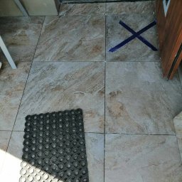 Dawid Szulik - Ułożone płytki podłogowe imitujące marmur, z niebieską taśmą w kształcie X na jednej z płytek i gumową wycieraczką w rogu, przy wejściu.