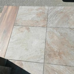 Dawid Szulik - Ułożone kwadratowe płyty tarasowe w odcieniach beżu i szarości, z widoczną strukturą kamienia, sąsiadujące z drewnopodobną płytą i szarą kostką brukową. W rogu widoczna wycieraczka.