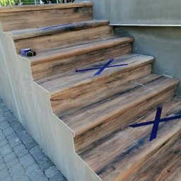 Dawid Szulik - Schody zewnętrzne wyłożone płytkami imitującymi drewno, zabezpieczone niebieską taśmą malarską, widoczny fragment chodnika z kostki brukowej i siedząca osoba w tle.