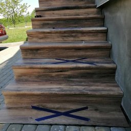Dawid Szulik - Schody zewnętrzne wyłożone płytkami imitującymi drewno, zabezpieczone taśmą malarską, z widoczną pacą do nakładania kleju.