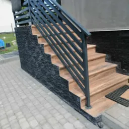 Zewnętrzne schody z okładziną z kamienia łupkowego i drewnopodobnymi stopniami, zabezpieczone nowoczesną, metalową balustradą w kolorze grafitowym, prowadzące z brukowanego chodnika.