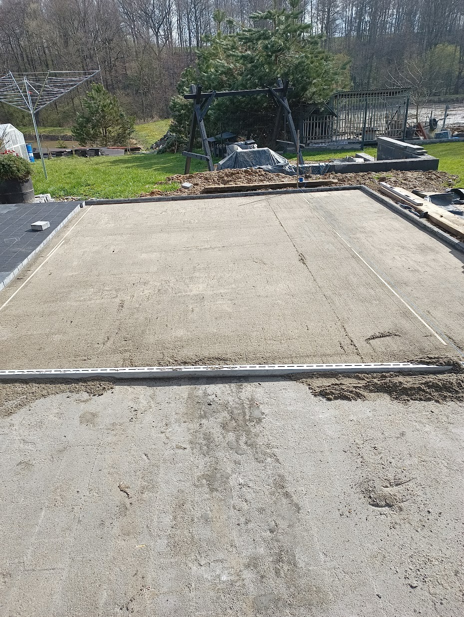 Przygotowany teren pod budowę tarasu z widocznymi profilami poziomującymi i piaskiem, w tle huśtawka ogrodowa i drzewa.