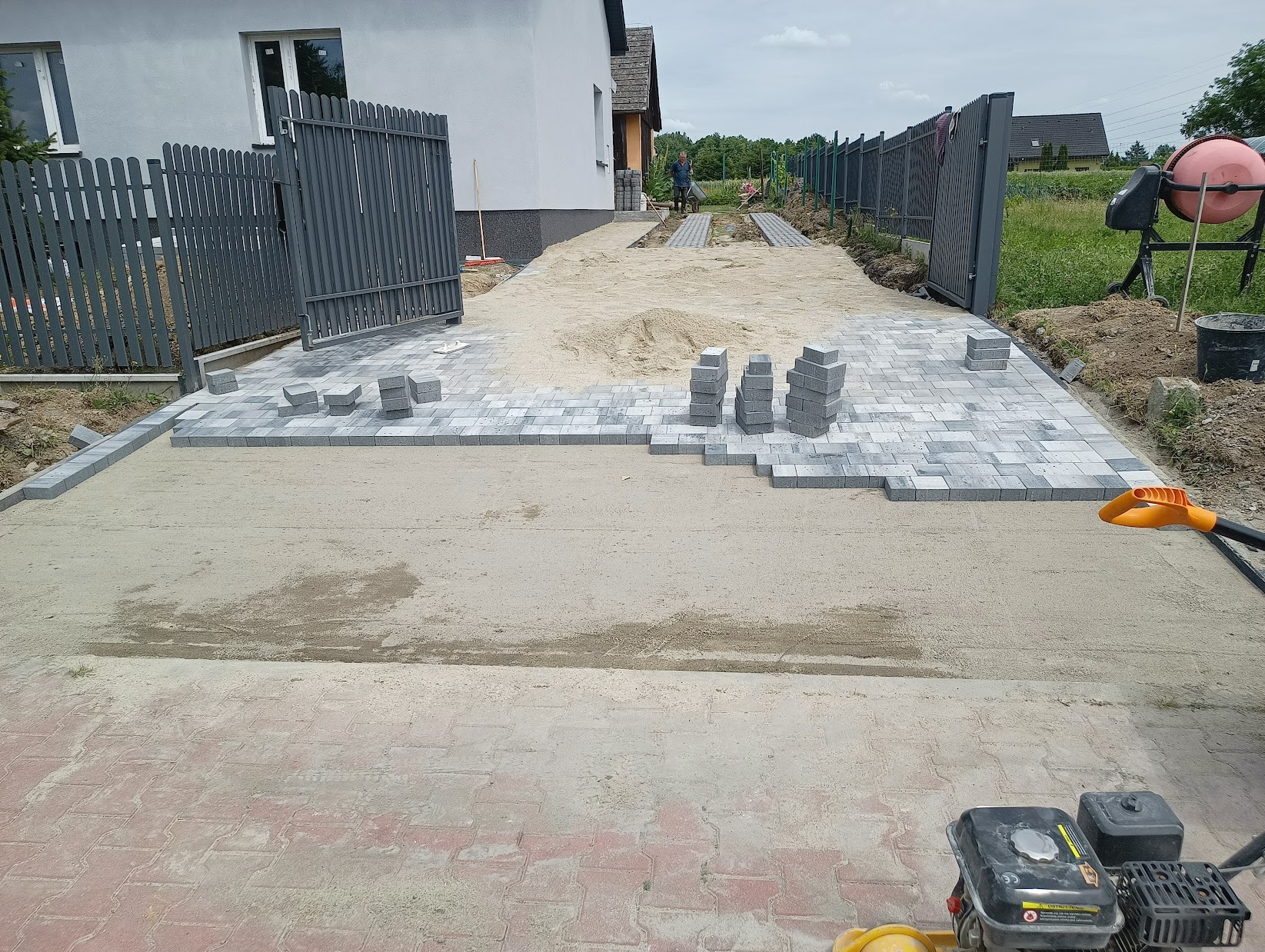 Ułożona częściowo szara kostka brukowa na podsypce piaskowej, widoczne stosy kostki, w tle dom z szarą elewacją, otwarte szare metalowe ogrodzenie i pracownik z taczką.