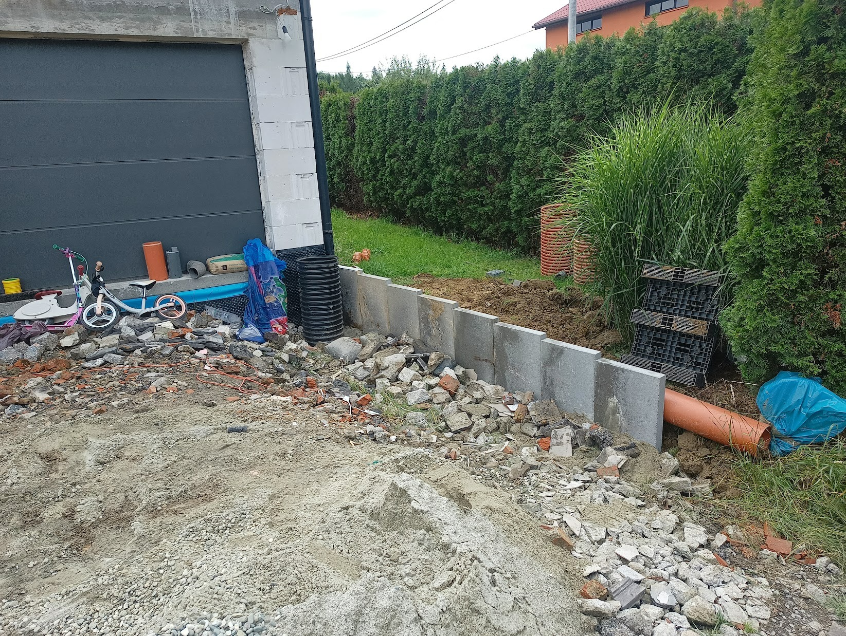 Przygotowanie podłoża pod kostkę brukową: wyrównany teren z widocznymi krawężnikami, gruzem i rurami drenażowymi, obok garażu i zieleni.