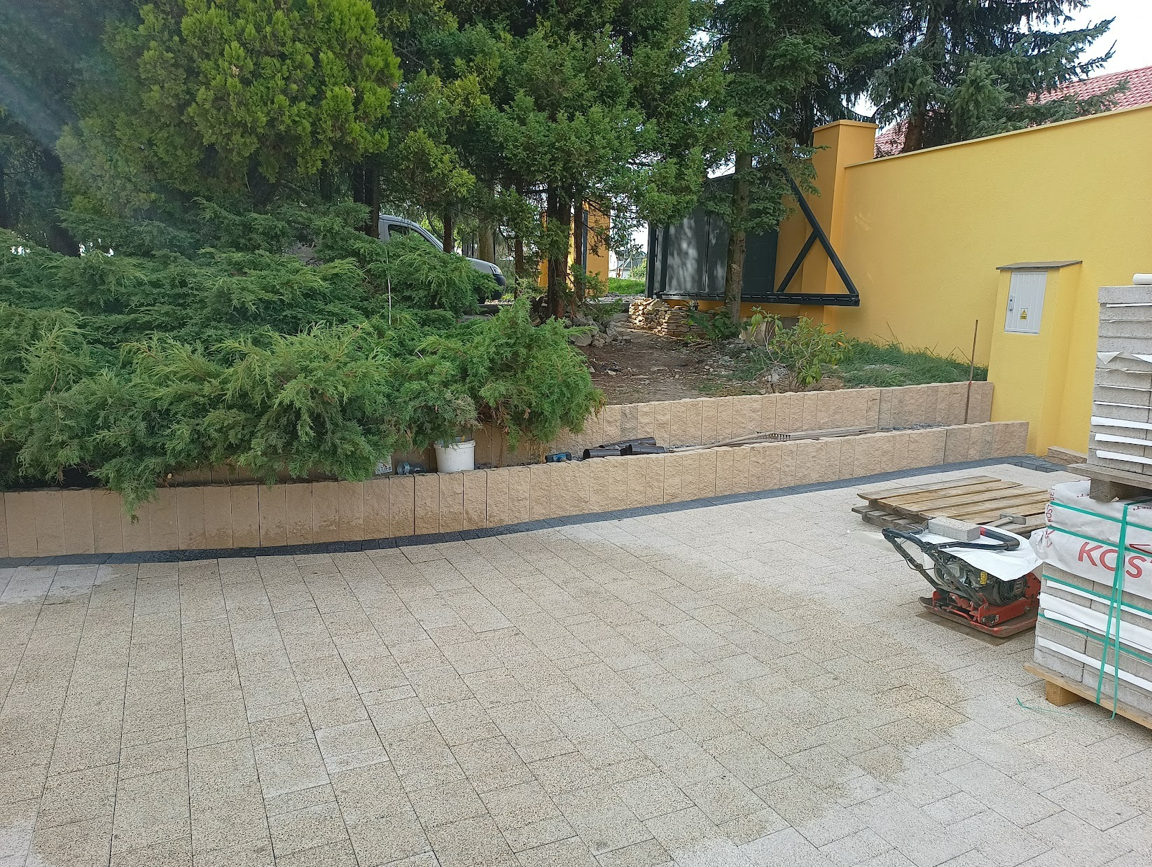 Ułożona jasna kostka brukowa na posesji z widoczną zagęszczarką, paletą kostki i podwyższonym rabatem z roślinami, na tle żółtej ściany budynku.