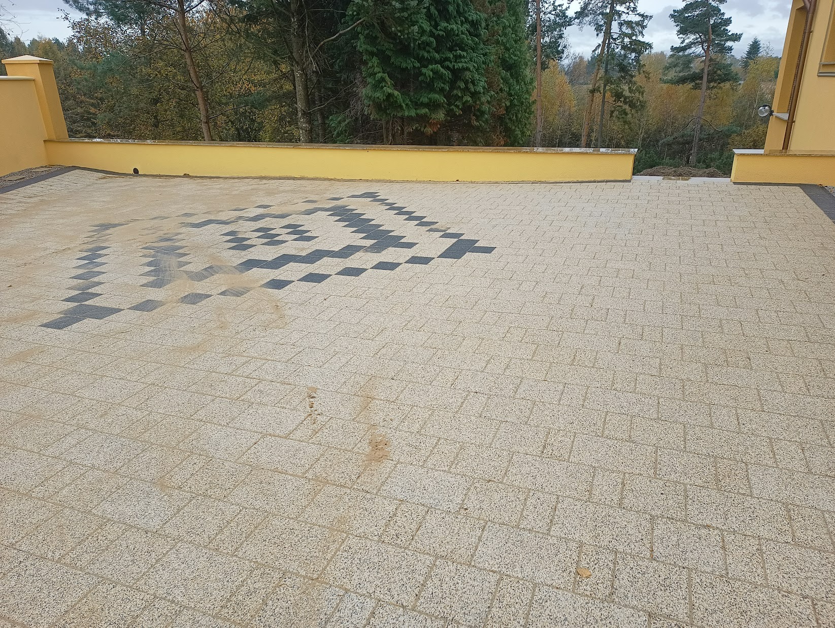 Ułożona kostka brukowa w geometryczny wzór z ciemnych i jasnych kostek na podjeździe, widoczne żółte ściany budynku i drzewa w tle.