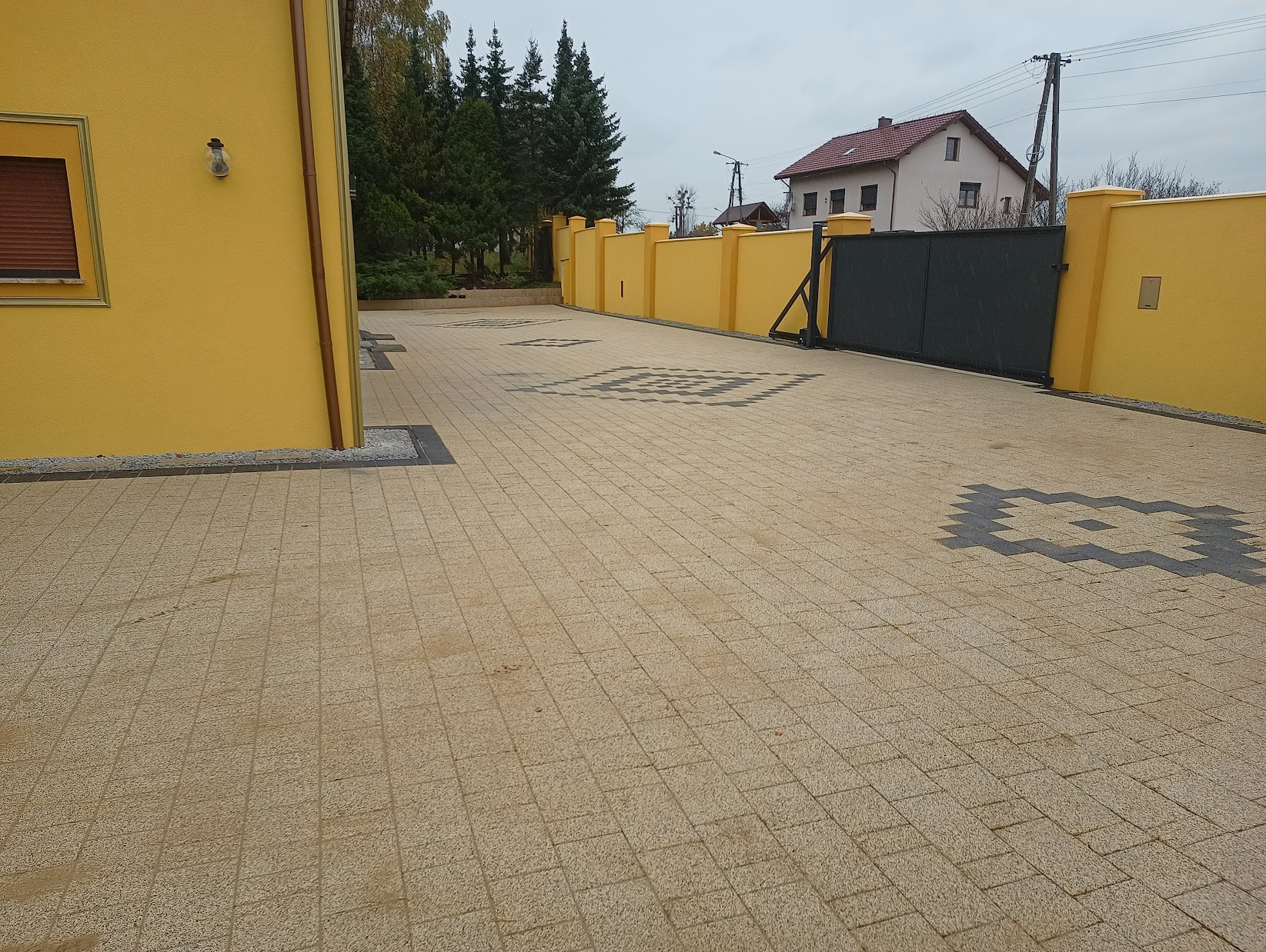 Beżowa kostka brukowa ułożona we wzór z ciemnymi akcentami, otoczona żółtym budynkiem i ogrodzeniem, z czarną bramą w tle.