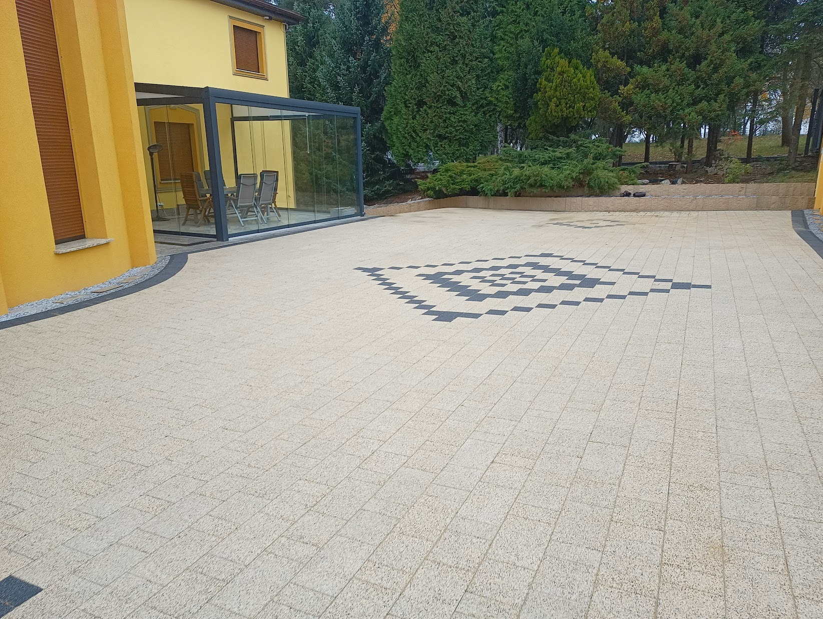 Jasna kostka brukowa ułożona we wzór szachownicy przed przeszkloną oranżerią przylegającą do żółtego budynku, otoczona zielenią.