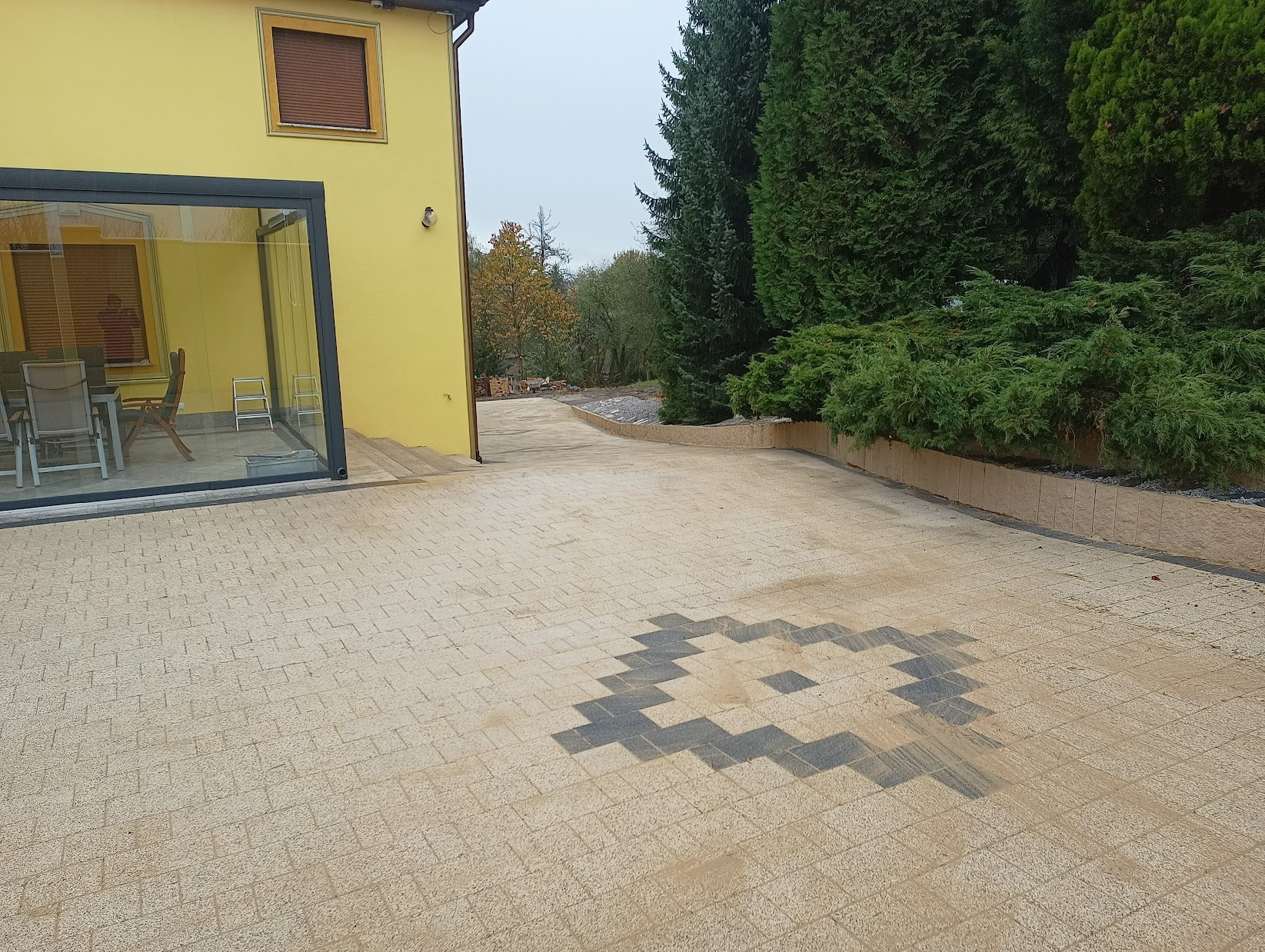 Beżowa kostka brukowa ułożona w geometryczny wzór na podjeździe, z elementami z ciemniejszej kostki tworzącymi dekorację. Żółty budynek z przeszklonym tarasem i zielone tuje w tle.