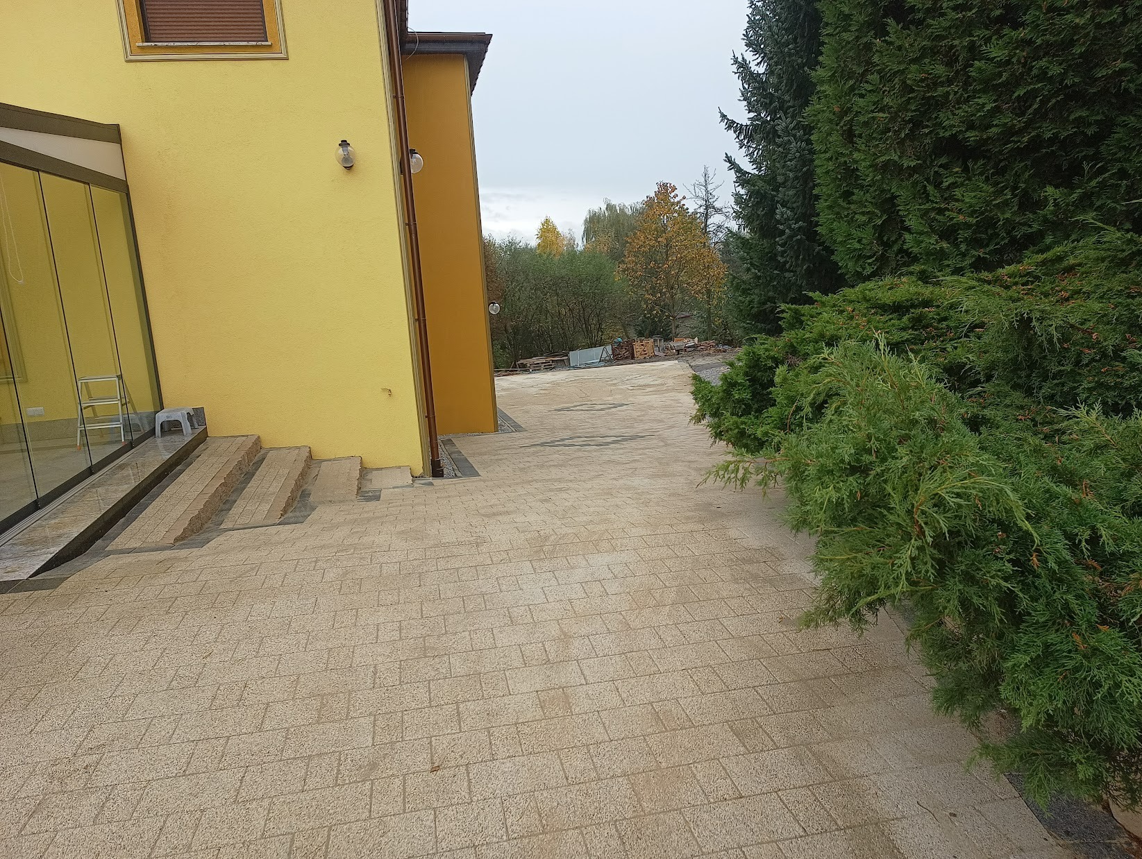 Beżowa kostka brukowa ułożona przed żółtym domem z przeszkloną werandą, widoczna zieleń drzew i krzewów w tle.