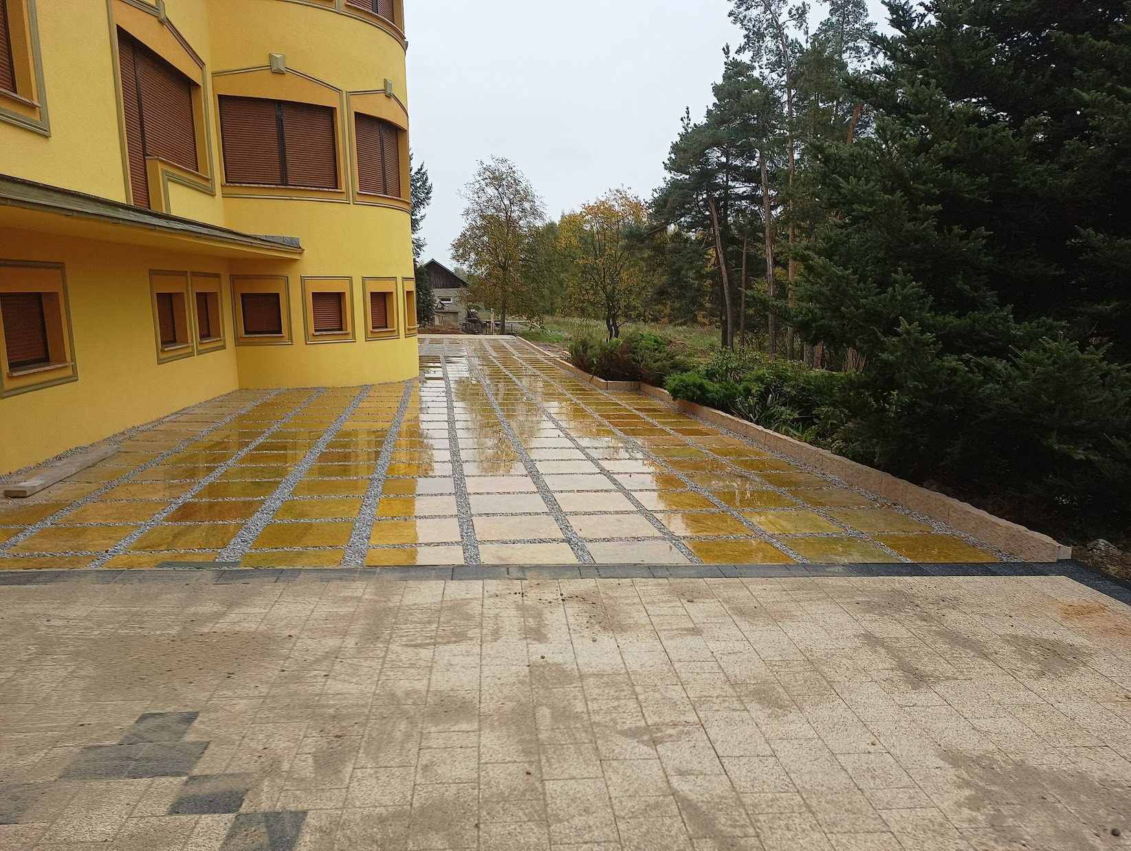 Żółta kostka brukowa ułożona w kwadraty z drobnym żwirem między nimi, prowadząca od budynku w kierunku drzew, po deszczu.