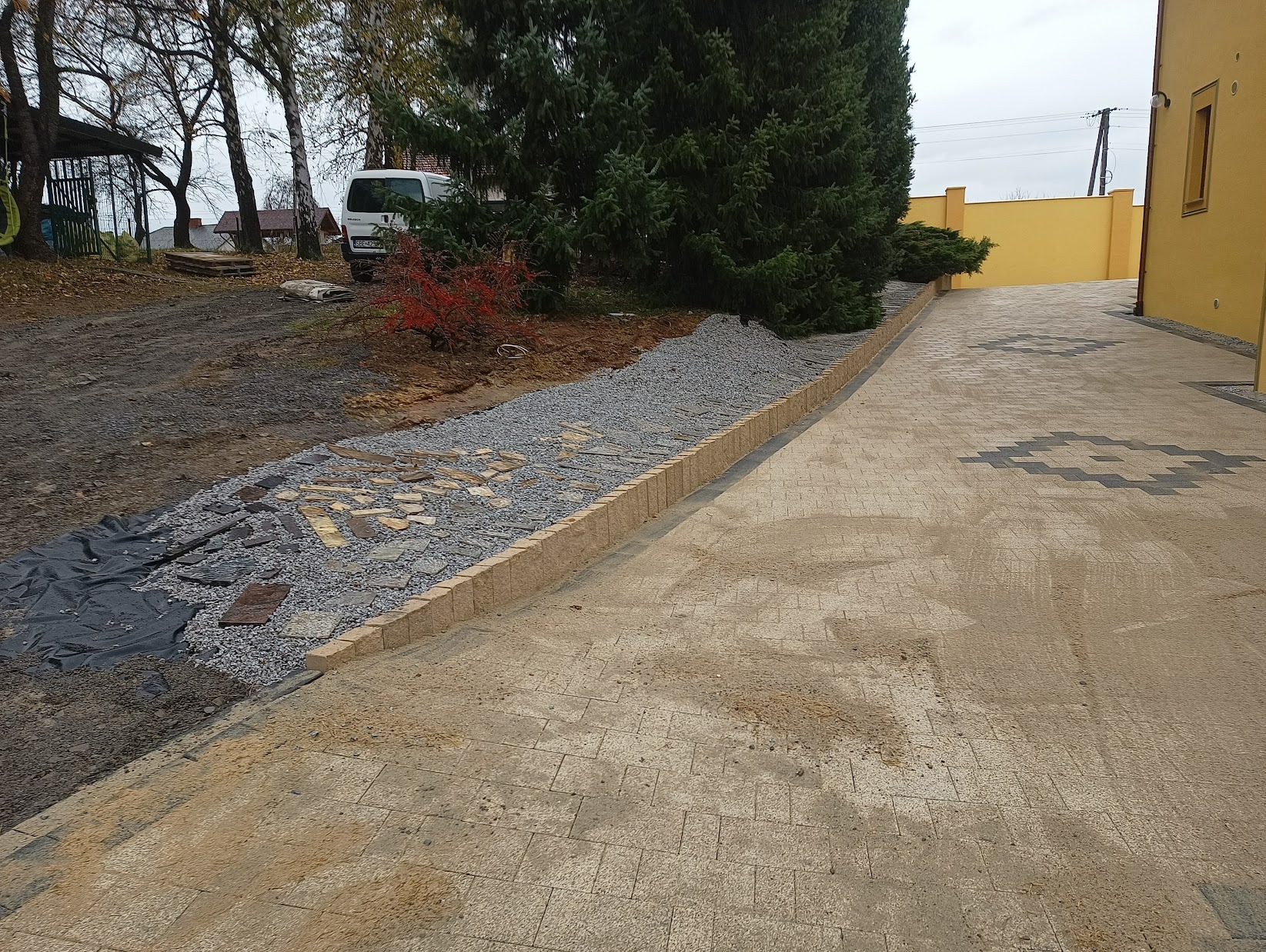 Ułożona kostka brukowa w geometryczny wzór, obok rabata z kamieniami i fragmentem czarnej geowłókniny, widoczny samochód dostawczy w tle.