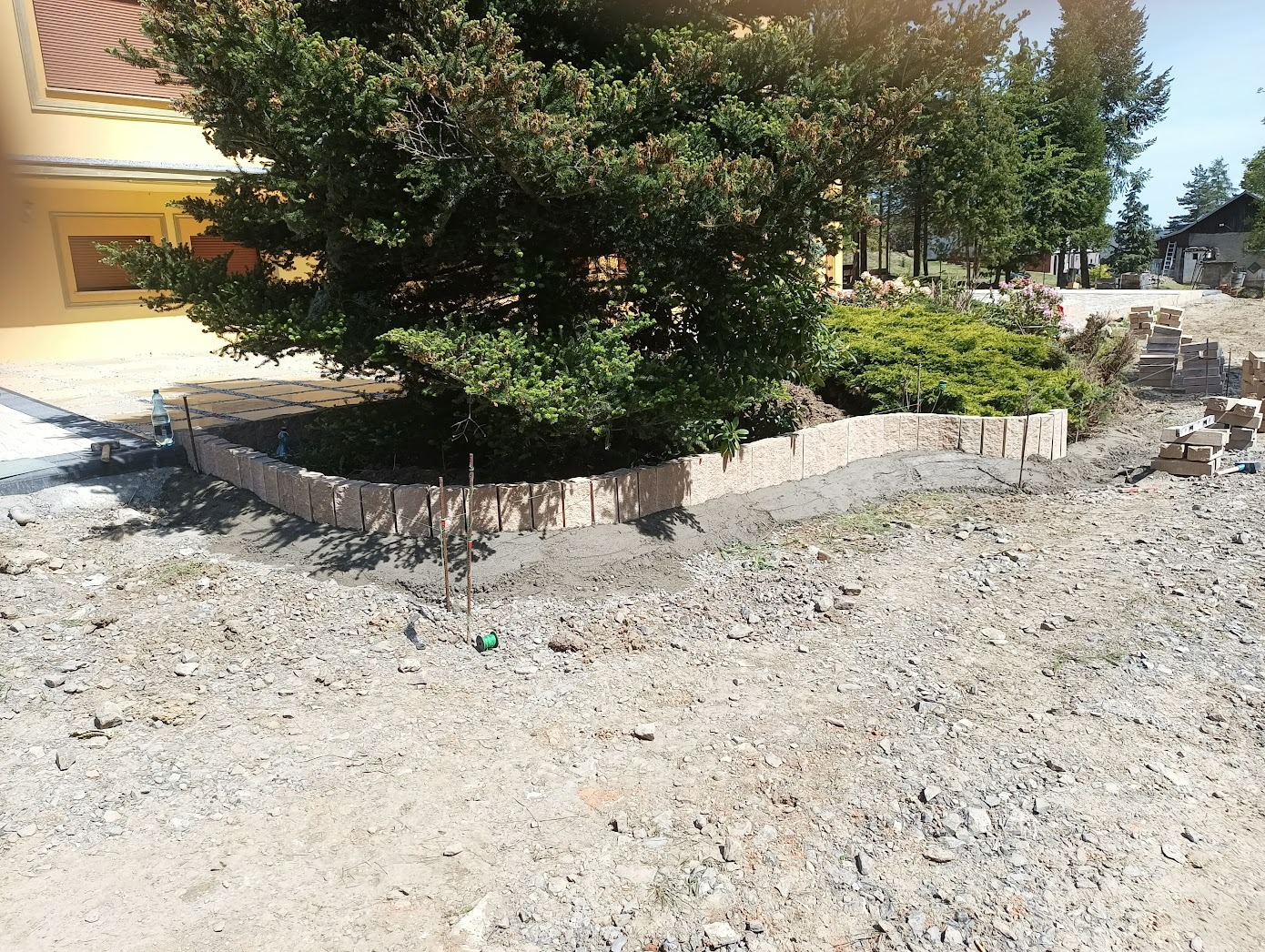 Budowa obrzeża rabaty z kostki, na podsypce cementowej, z widocznymi palikami wyznaczającymi łuk, drzewo w centralnej części rabaty, materiał budowlany składowany obok.