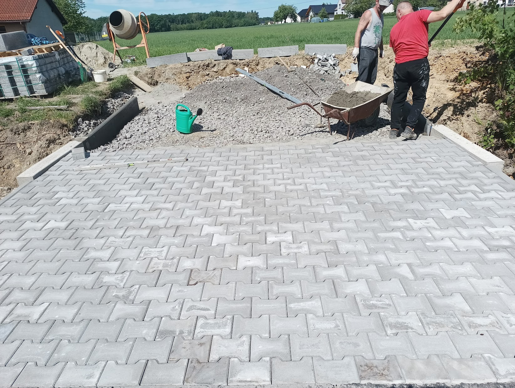 Ułożona szara kostka brukowa na posesji, w tle pracownicy z taczką, betoniarka i widok na zielone pole oraz zabudowania.